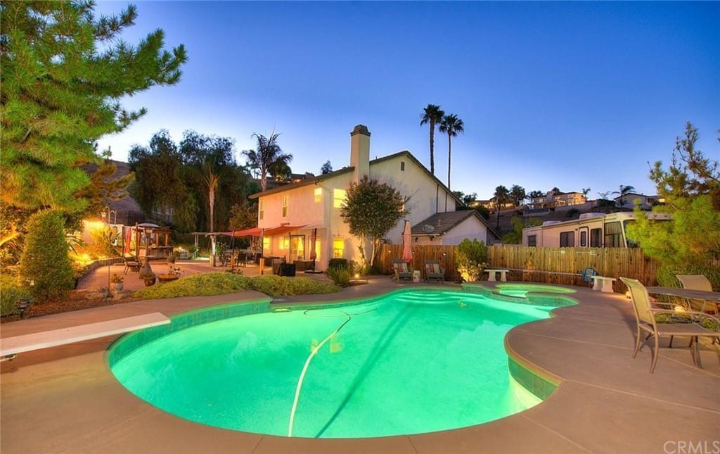Backyard oasis featuring lit pool and large house with trees and concrete patio
